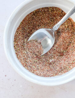 A silver spoon rests in a small white bowl filled with homemade spice mix, a reddish-brown spice blend speckled with green and yellow herbs, on a light-colored surface.