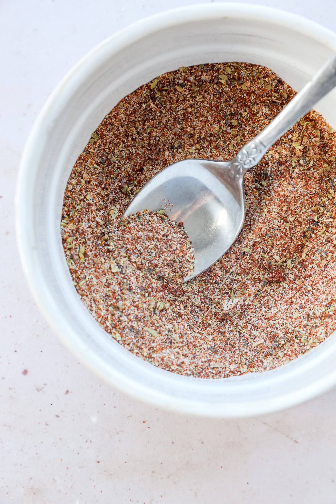 A silver spoon rests in a small white bowl filled with homemade spice mix, a reddish-brown spice blend speckled with green and yellow herbs, on a light-colored surface.