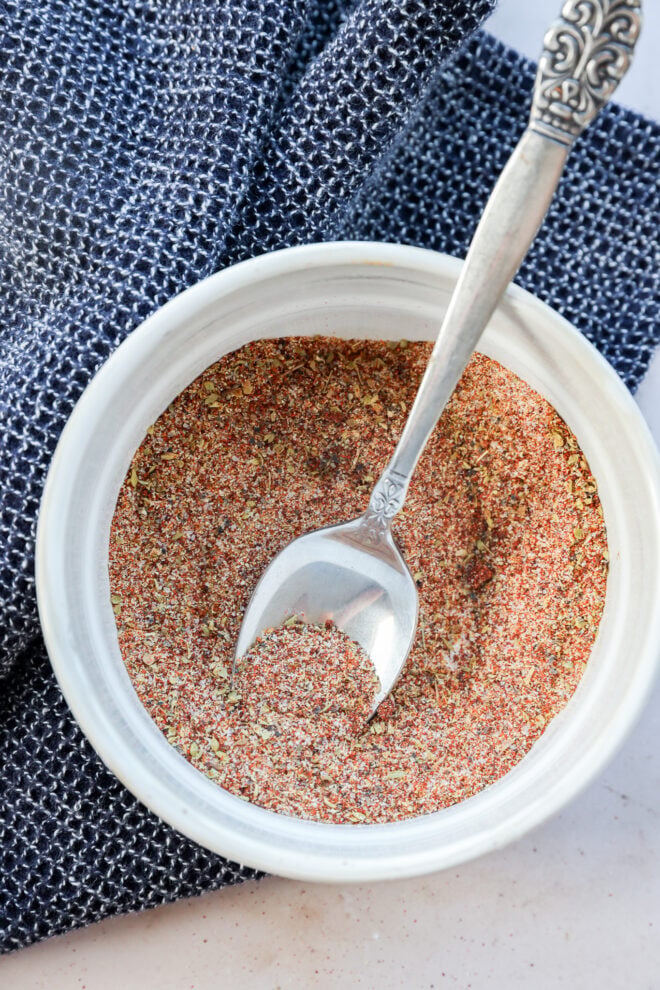 A small white bowl filled with a reddish-brown Carne Asada Seasoning mix sits on a blue textured cloth, with a silver spoon resting inside the bowl.
