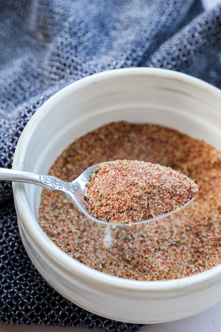A spoonful of Carne Asada Seasoning is held over a white bowl filled with the same spice blend, with a textured dark blue cloth in the background.