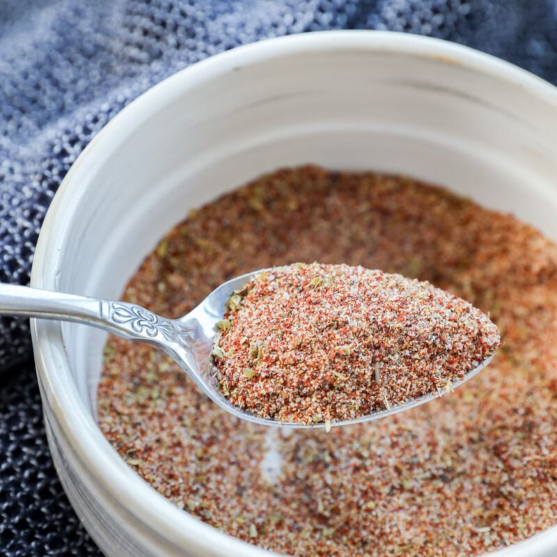 A spoonful of Carne Asada Seasoning is held over a white bowl filled with the same spice blend, with a textured dark blue cloth in the background.