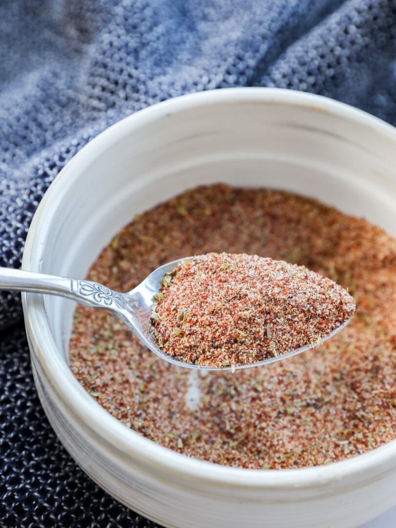 A spoonful of Carne Asada Seasoning is held over a white bowl filled with the same spice blend, with a textured dark blue cloth in the background.