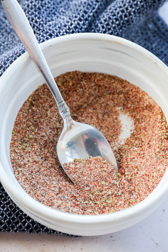 A white bowl filled with a flavorful spice blend and a silver spoon rests inside, atop a textured blue cloth.