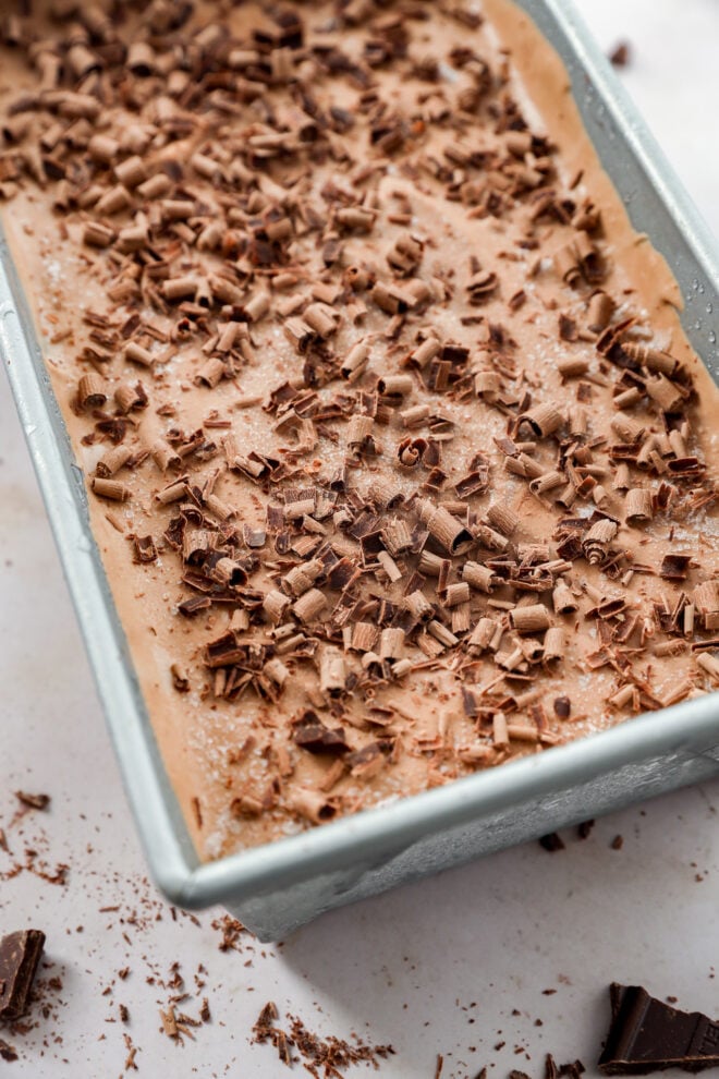 A close-up of a loaf pan filled with creamy Death By Chocolate Ice Cream, topped with chocolate shavings and surrounded by small pieces of chocolate on a light surface.