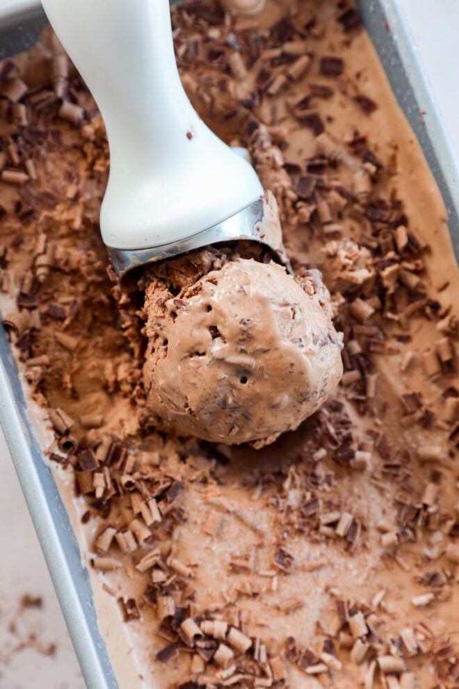A metal ice cream scoop holds a serving of decadent Death By Chocolate Ice Cream with chocolate shavings on top, resting in a pan filled with more chocolate ice cream and shavings.