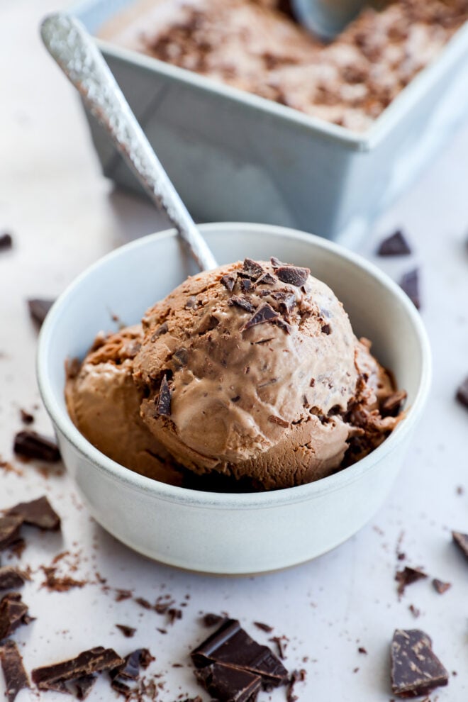 A bowl with chocolate chunks and a spoon, with more chocolate pieces and ice cream in a container in the background.