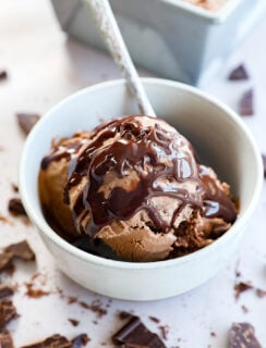 A white bowl filled with Death By Chocolate Ice Cream topped with rich chocolate syrup, with a spoon inside. Broken chocolate pieces are scattered around the bowl on a light surface.