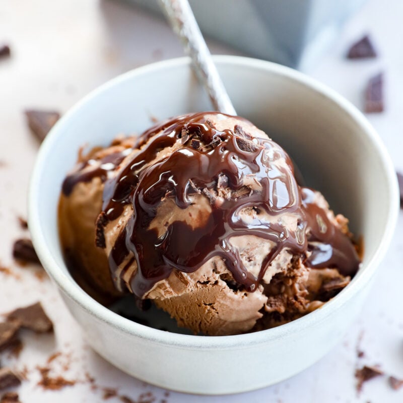 A white bowl filled with Death By Chocolate Ice Cream topped with rich chocolate syrup, with a spoon inside. Broken chocolate pieces are scattered around the bowl on a light surface.