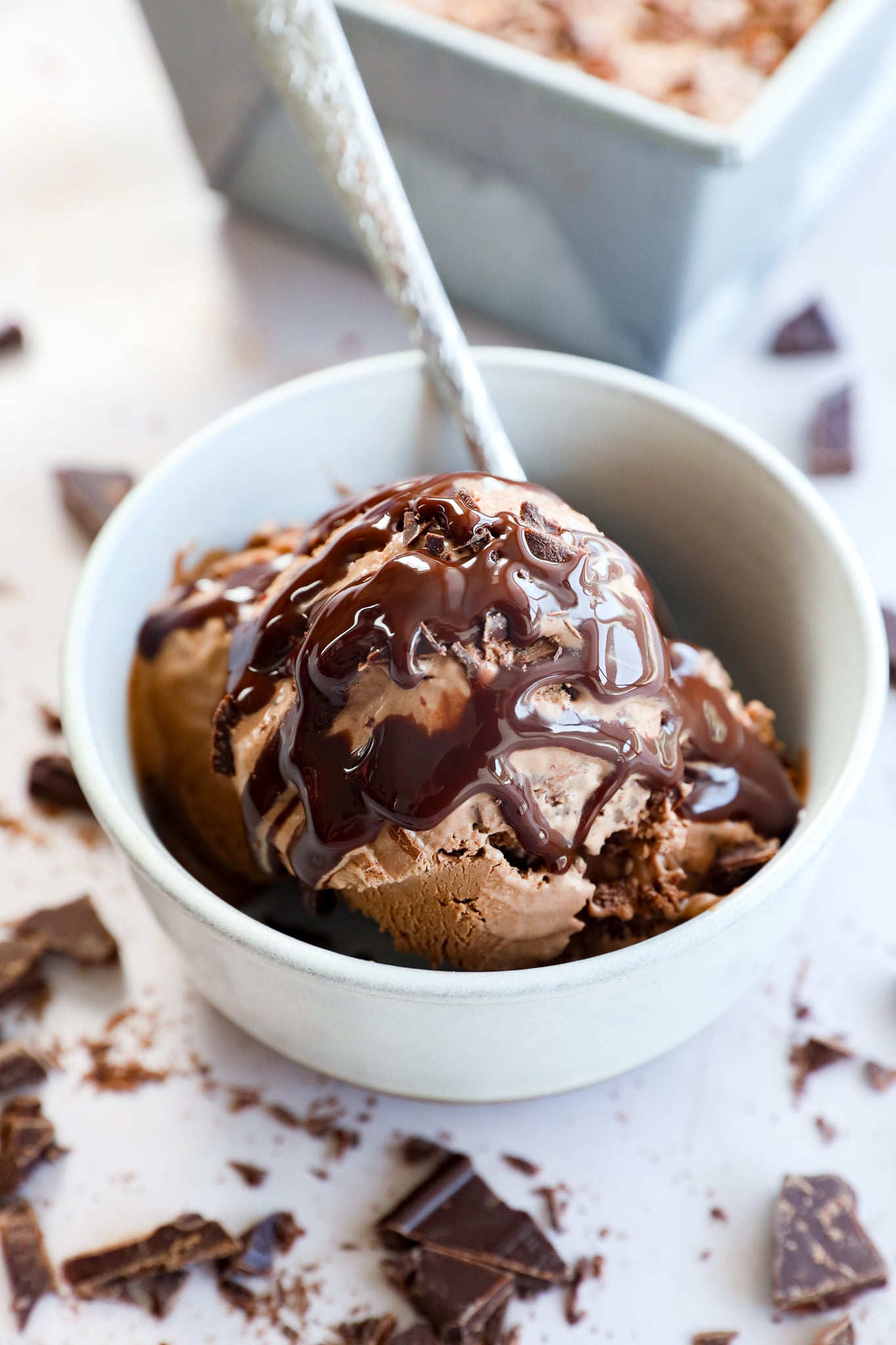 A white bowl filled with Death By Chocolate Ice Cream topped with rich chocolate syrup, with a spoon inside. Broken chocolate pieces are scattered around the bowl on a light surface.