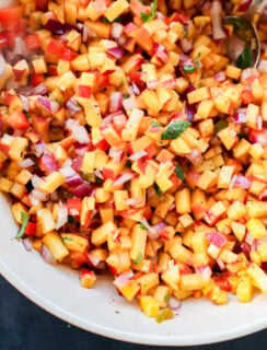 A close-up of a bowl filled with diced peaches, red onions, red bell peppers, and chopped herbs, all mixed together, with a spoon visible in the top right corner.