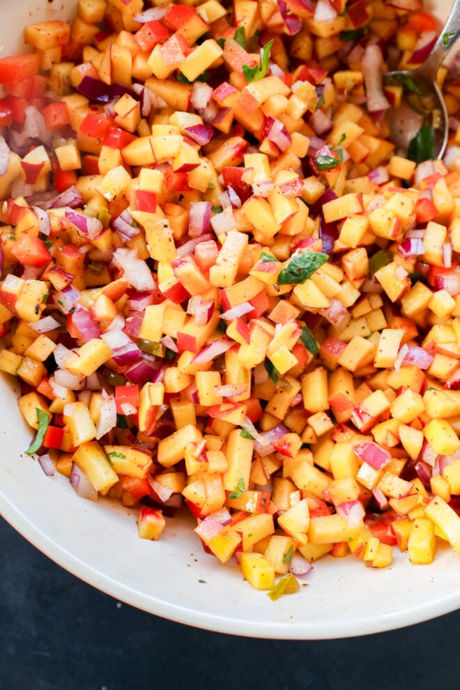 A close-up of a bowl filled with diced peaches, red onions, red bell peppers, and chopped herbs, all mixed together, with a spoon visible in the top right corner.