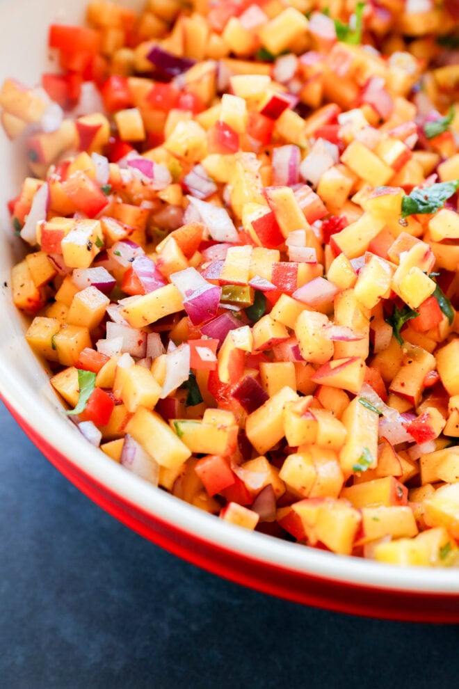 A close-up of a bowl filled with colorful diced fruits and vegetables, including peaches, red onions, red peppers, and fresh herbs.