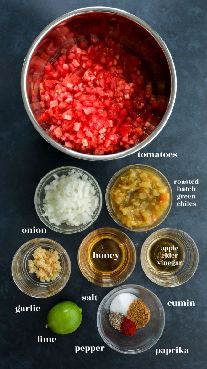 Overhead view of bowls containing ingredients for tomato chutney: chopped tomatoes, onions, garlic, lime, roasted hatch green chiles, honey, apple cider vinegar, salt, pepper, cumin, and paprika on a dark surface.