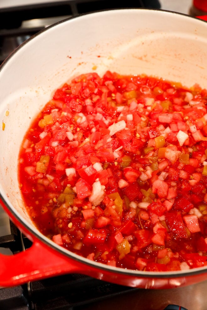 A close-up of a red pot on a stove filled with chopped tomatoes, onions, and green peppers simmering in liquid, likely being cooked into a flavorful tomato chutney or sauce.