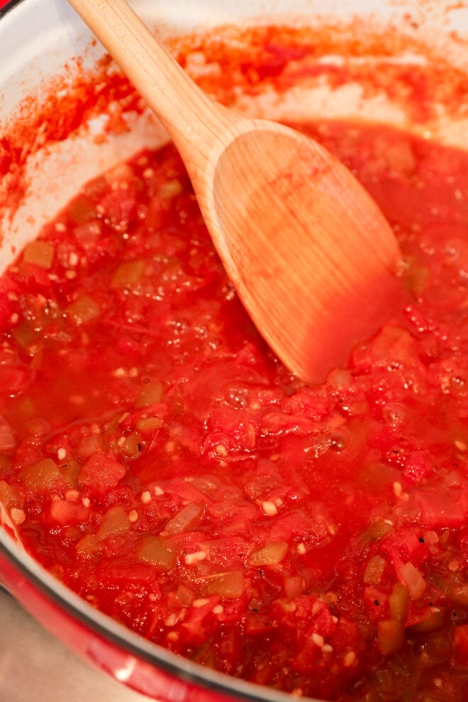 A wooden spoon stirs a pot of chunky tomato chutney with visible pieces of tomato, onion, and garlic.