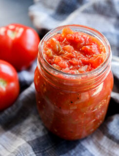 A glass jar filled with homemade tomato sauce sits on a plaid cloth, accompanied by two fresh tomatoes in the background, evoking the vibrant flavors of tomato chutney.