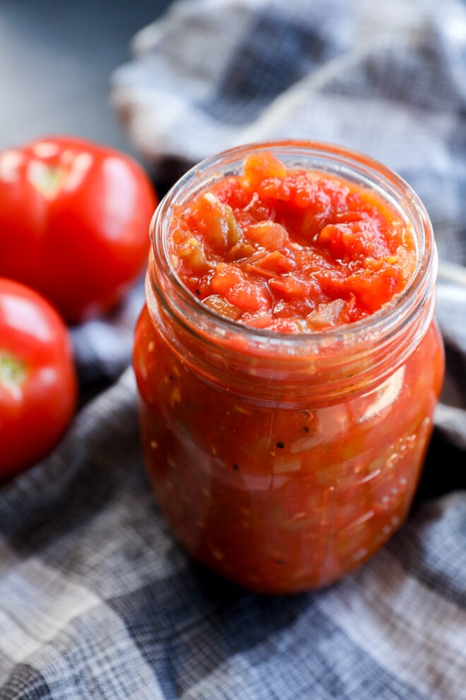A glass jar filled with homemade tomato sauce sits on a plaid cloth, accompanied by two fresh tomatoes in the background, evoking the vibrant flavors of tomato chutney.