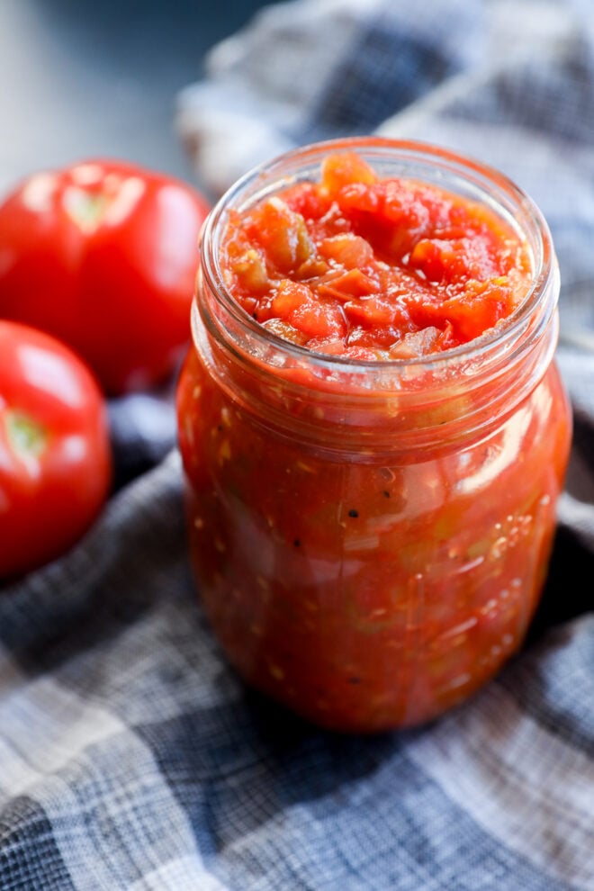 A glass jar filled with chunky tomato chutney sits on a plaid cloth, with two fresh tomatoes in the background.