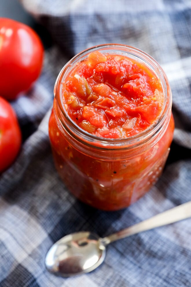 A glass jar filled with chunky hatch green chile sauce sits on a blue plaid cloth, next to fresh tomatoes and a silver spoon.