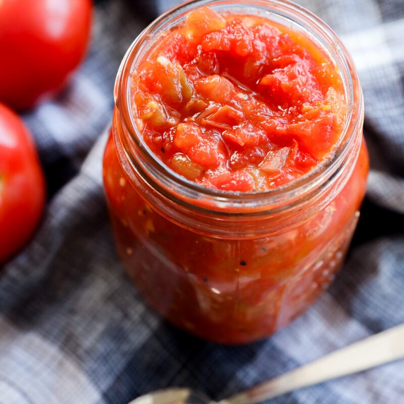 A glass jar filled with chunky hatch green chile sauce sits on a blue plaid cloth, next to fresh tomatoes and a silver spoon.