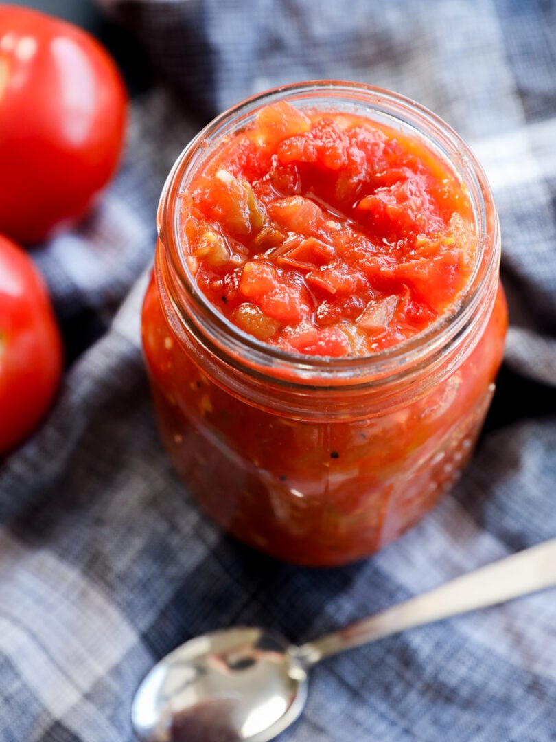 A glass jar filled with chunky hatch green chile sauce sits on a blue plaid cloth, next to fresh tomatoes and a silver spoon.