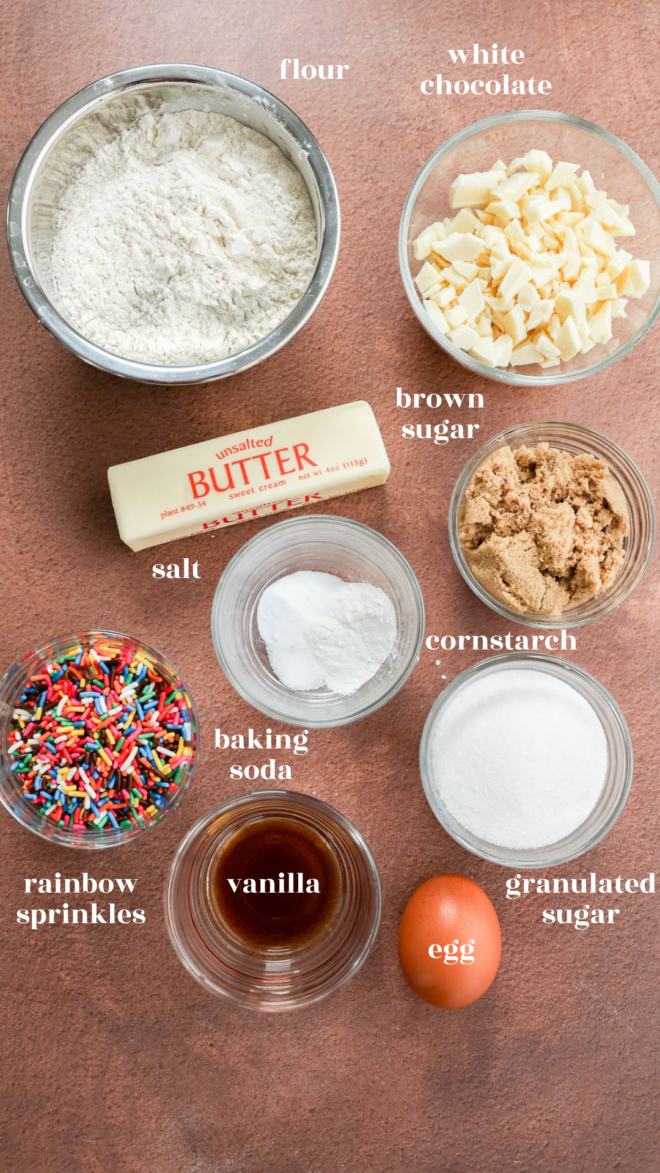 Baking ingredients for birthday cake cookies—including flour, white chocolate chunks, butter, brown sugar, salt, baking soda, cornstarch, rainbow sprinkles, vanilla, granulated sugar, and an egg—are arranged in bowls on a brown surface.