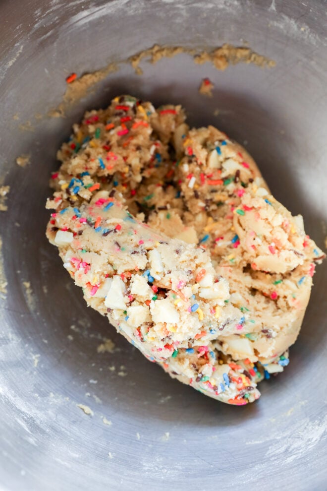 A close-up of raw cookie dough with colorful sprinkles and white chocolate chunks in a metal mixing bowl.