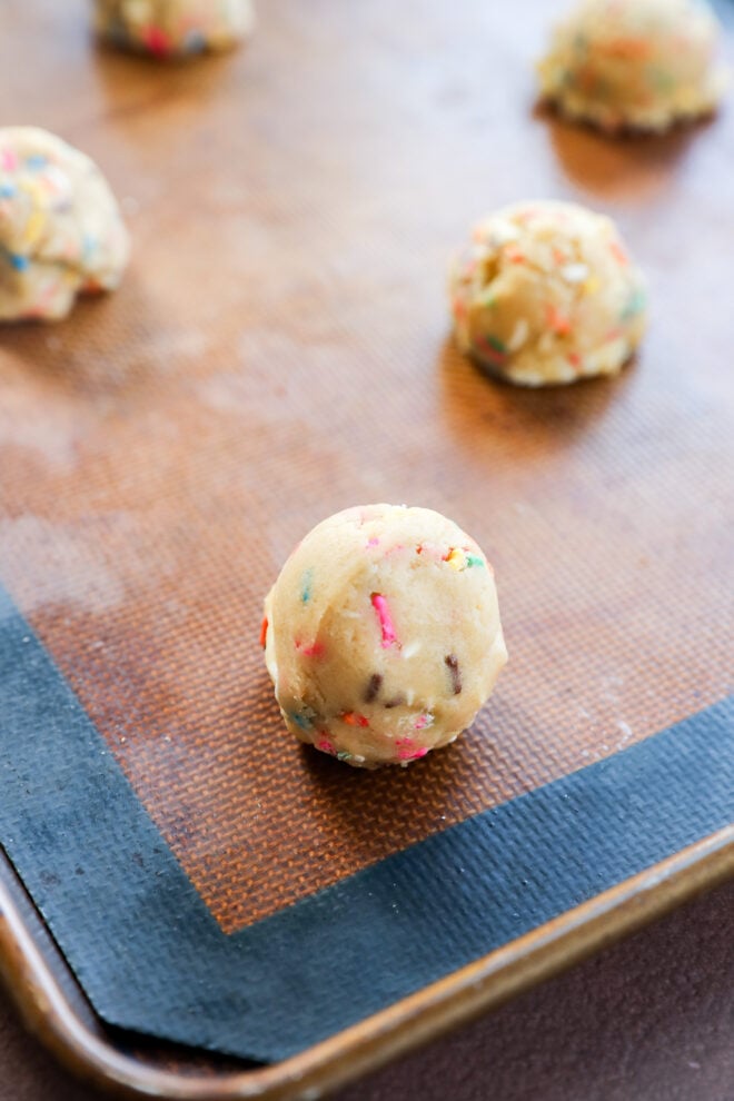 Close-up of round birthday cake cookies dough balls with colorful sprinkles on a silicone baking mat, placed on a metal baking sheet, ready to be baked.