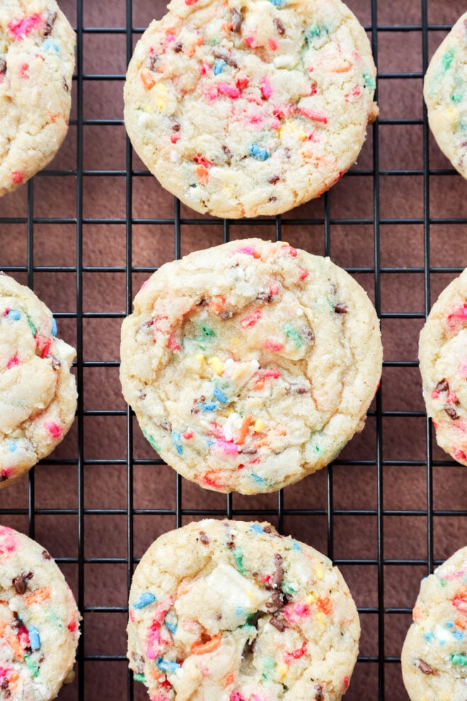 Birthday cake cookies with colorful sprinkles are cooling on a black wire rack over a brown surface, arranged in neat rows.
