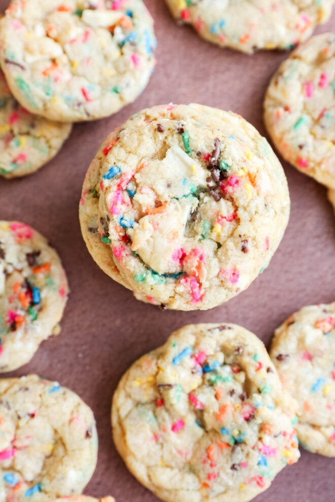 Close-up of several colorful birthday cake cookies on a brown surface, showing a crumbly texture and rainbow sprinkles baked throughout the soft, round cookies.