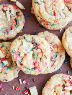 A close-up of colorful dessert with rainbow sprinkles and chunks of white chocolate, some stacked and some scattered on a brown surface.
