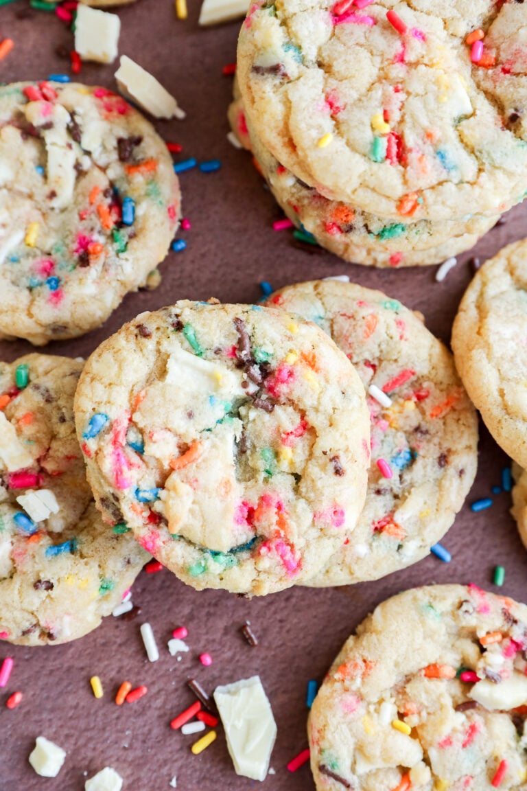 A close-up of colorful dessert with rainbow sprinkles and chunks of white chocolate, some stacked and some scattered on a brown surface.
