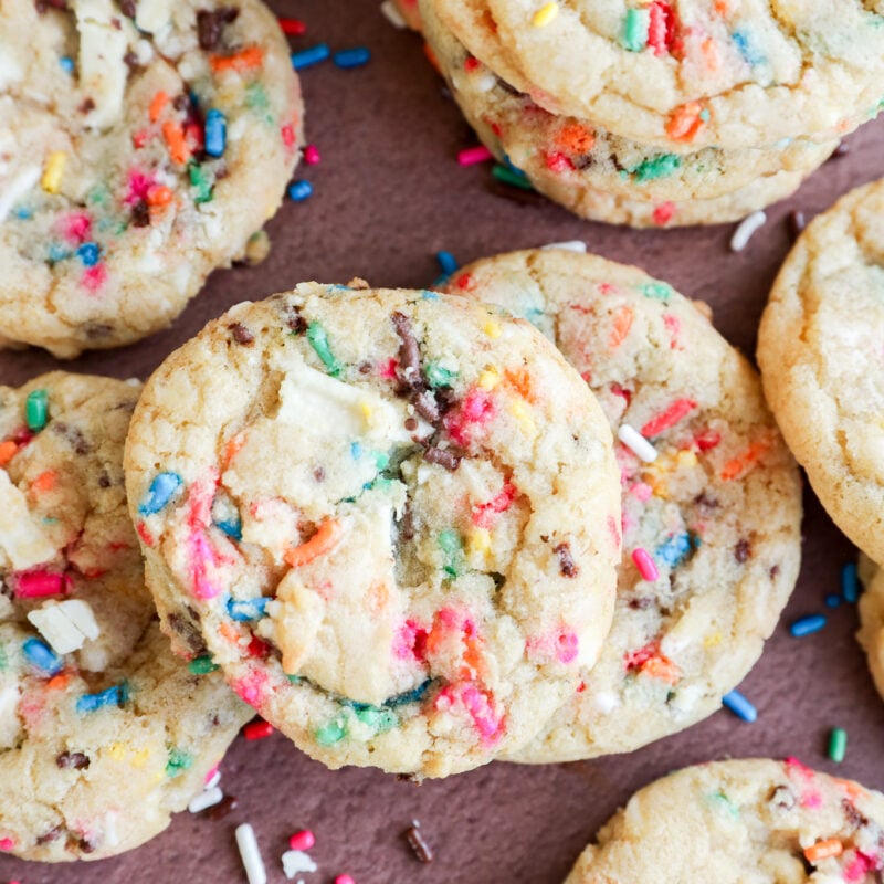 A close-up of colorful dessert with rainbow sprinkles and chunks of white chocolate, some stacked and some scattered on a brown surface.