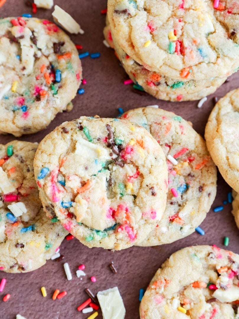 A close-up of colorful dessert with rainbow sprinkles and chunks of white chocolate, some stacked and some scattered on a brown surface.