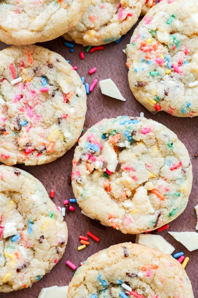 Close-up of birthday cake cookies with colorful rainbow sprinkles and white chocolate chunks scattered on a brown surface, with extra sprinkles and chocolate pieces around the cookies.