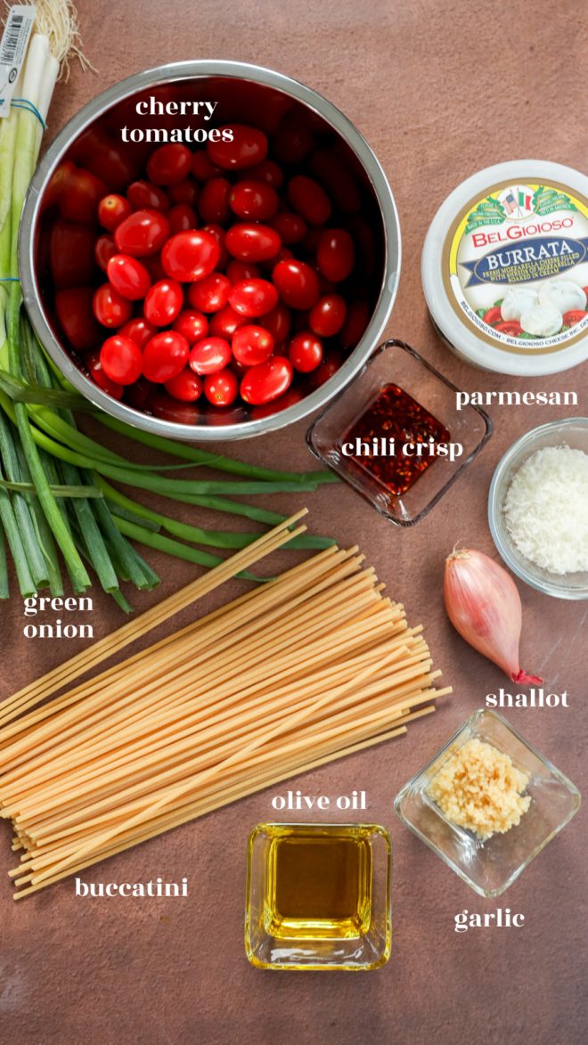 Ingredients for a Creamy Burrata Pasta with Chili Crisp & Burst Tomatoes, including cherry tomatoes, burrata, parmesan, chili crisp, green onion, shallot, bucatini pasta, olive oil, and minced garlic, are arranged neatly on a countertop. Labels identify each ingredient.