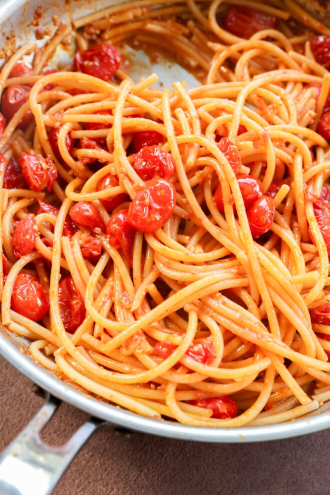 Buccatini in a pan, beautifully mixed with cherry tomatoes and tomato sauce, with a shiny metal handle visible in the lower left corner.