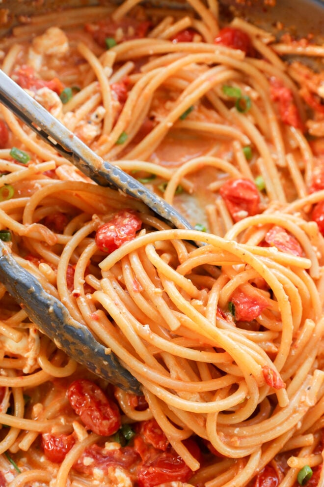 Tongs holding a serving of buccatini with cherry tomatoes visible in the pan, all coated in a rich sauce.