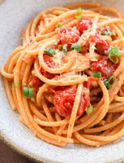 A bowl of Creamy Burrata Pasta with Chili Crisp & Burst Tomatoes, featuring cherry tomatoes, grated cheese, and chopped green onions, served in a rustic white bowl.
