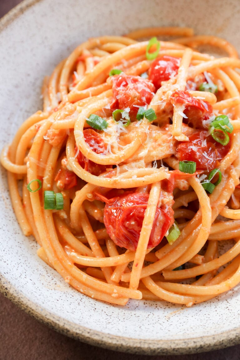 A bowl of Creamy Burrata Pasta with Chili Crisp & Burst Tomatoes, featuring cherry tomatoes, grated cheese, and chopped green onions, served in a rustic white bowl.