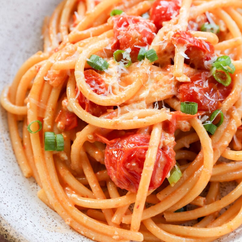 A bowl of Creamy Burrata Pasta with Chili Crisp & Burst Tomatoes, featuring cherry tomatoes, grated cheese, and chopped green onions, served in a rustic white bowl.