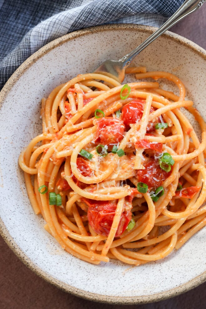 A bowl of Creamy Burrata Pasta with Chili Crisp & Burst Tomatoes, topped with grated cheese and chopped green onions, is served with a fork on a rustic plate and accompanied by a cloth napkin nearby.