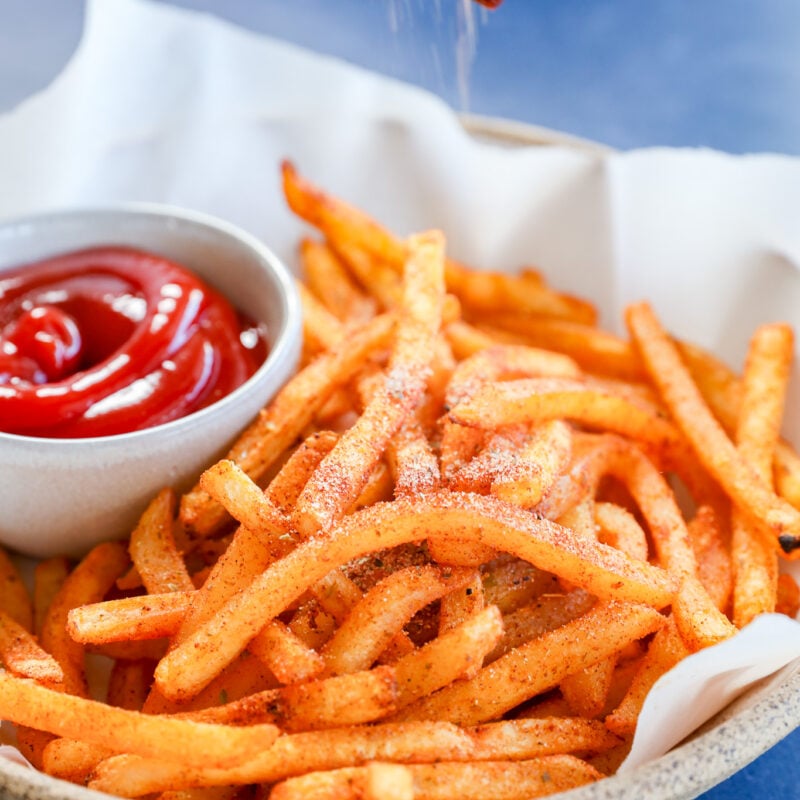 A hand sprinkles French Fry Seasoning over a plate of seasoned French fries with a small bowl of ketchup on the side, set on white parchment paper and a blue surface.