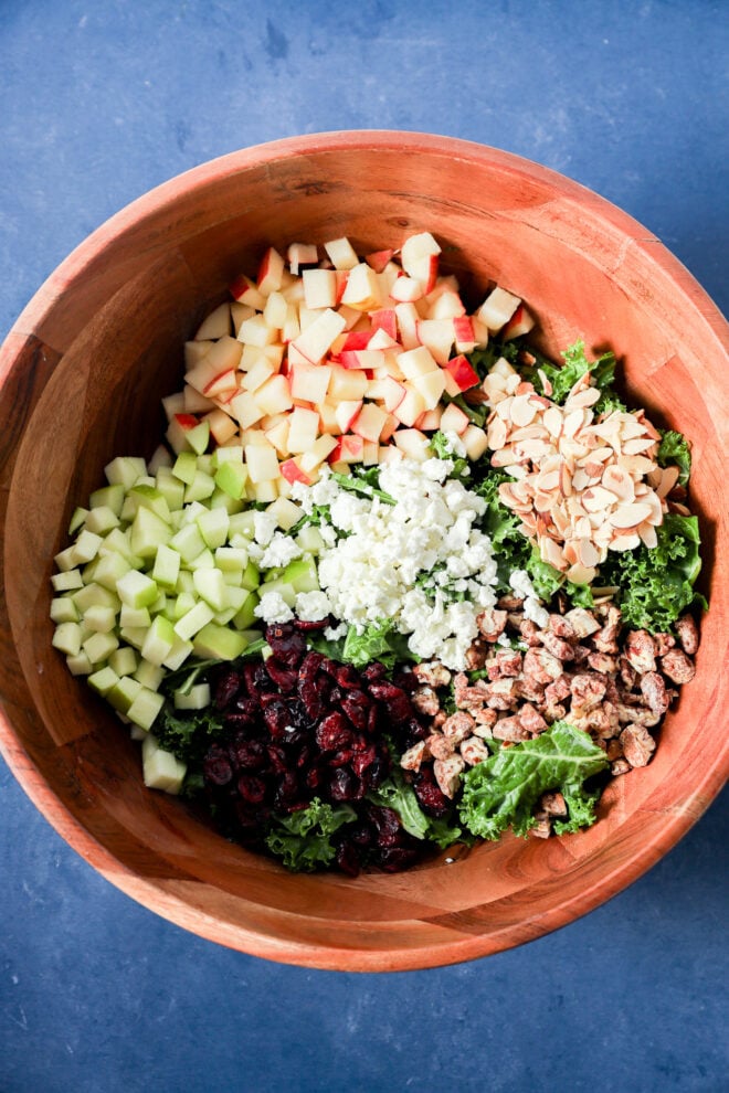 A wooden bowl filled with an apple and kale salad topped with diced red and green apples, crumbled cheese, dried cranberries, sliced almonds, and candied pecans.