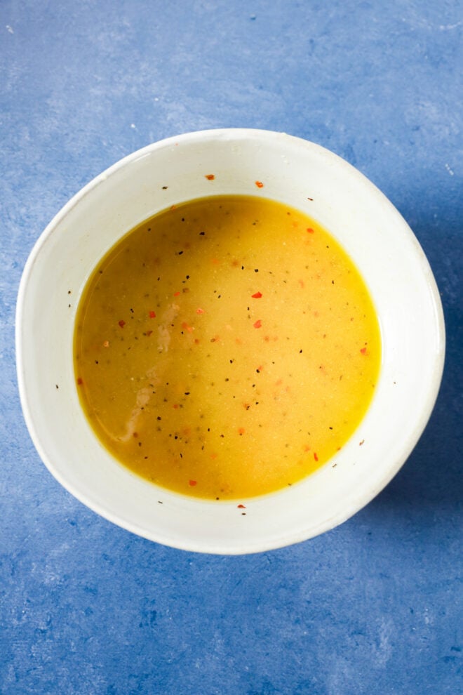 A white bowl filled with golden yellow dressing, seasoned with black pepper and red pepper flakes.