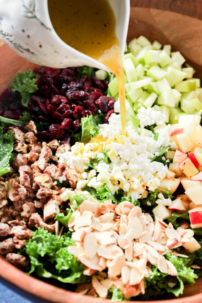 Vinaigrette being poured from a pitcher onto an apple and kale salad of dried cranberries, chopped apples, feta cheese, nuts, and sliced almonds in a wooden bowl.