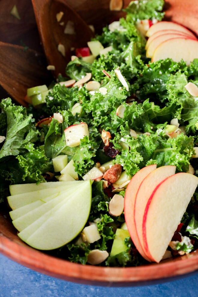 Apple and kale salad with dried cranberries and slivered almonds in a wooden bowl with serving spoons.
