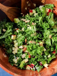 Apple and kale salad with chopped apples, dried cranberries, and sliced almonds in a wooden bowl with serving spoons.