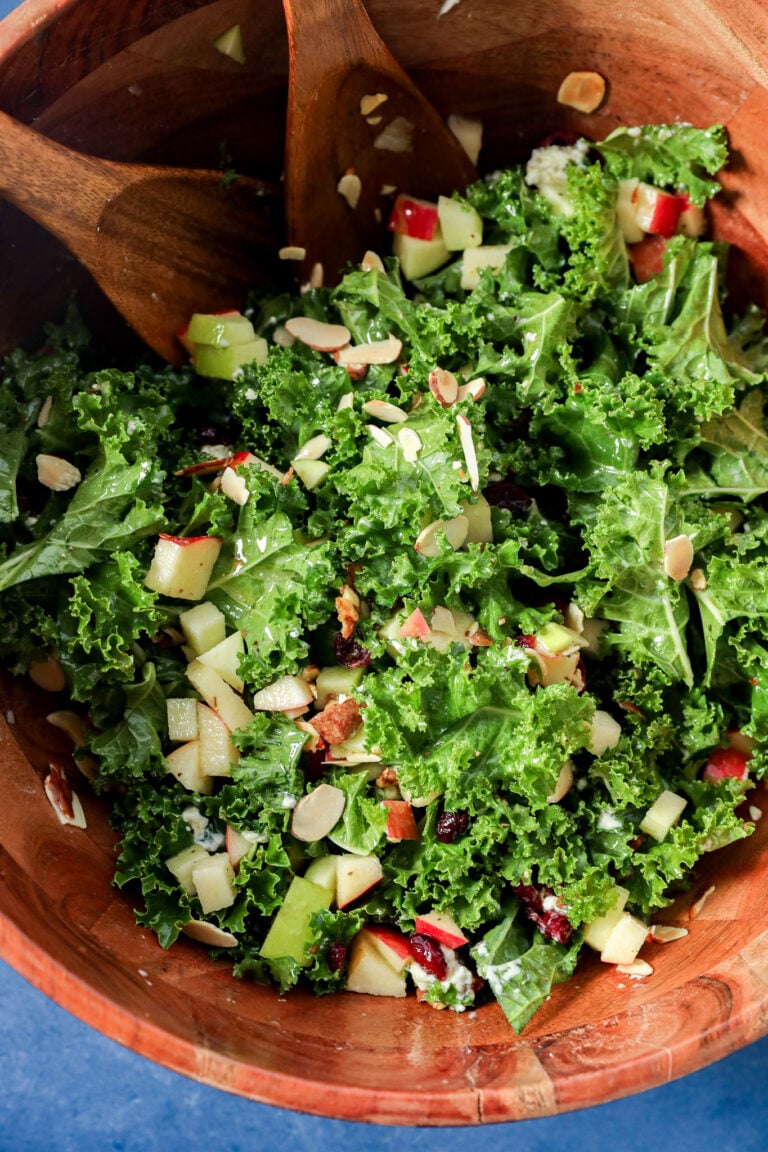 Apple and kale salad with chopped apples, dried cranberries, and sliced almonds in a wooden bowl with serving spoons.