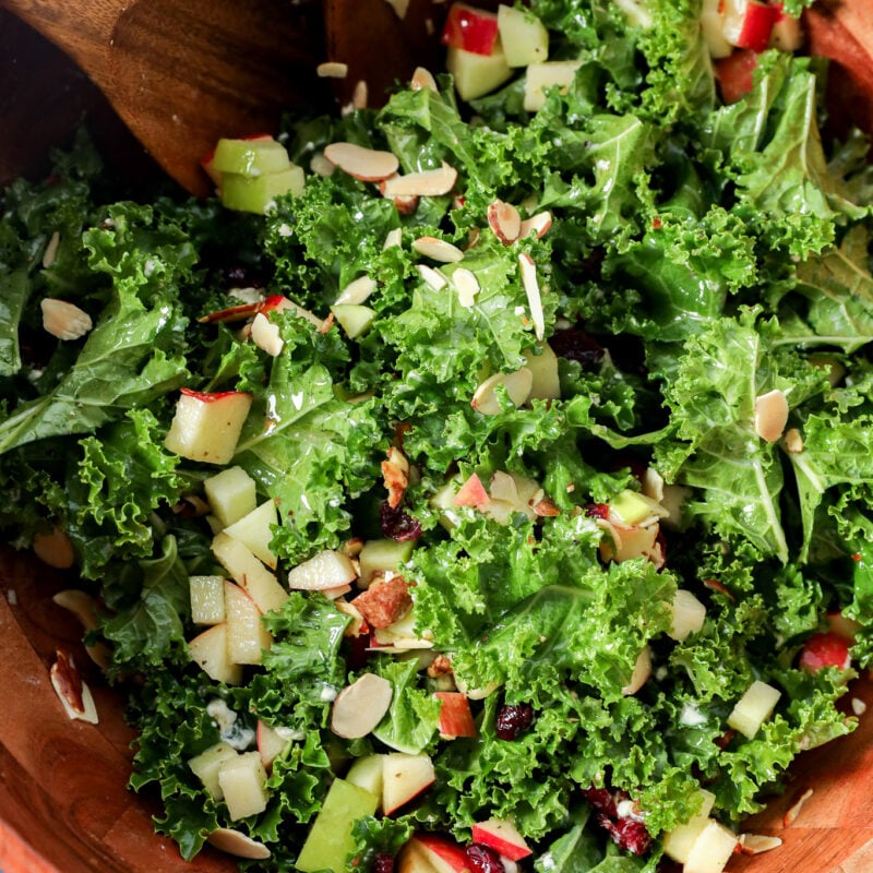 Apple and kale salad with chopped apples, dried cranberries, and sliced almonds in a wooden bowl with serving spoons.